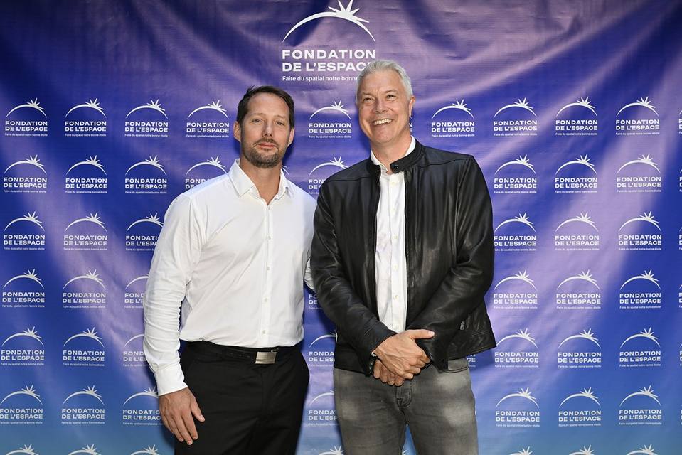 Deux convives, dont l'un en veste de cuir, devant le mur aux logos de la Fondation